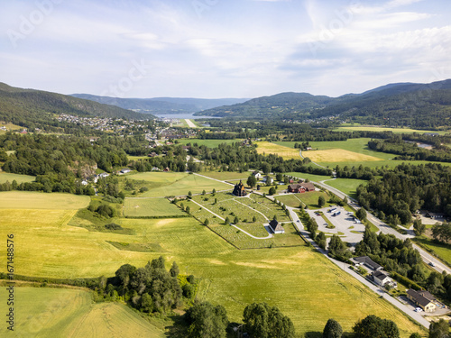 Aerial view of the Heddal Stave Church, a dark wooden structure amidst a green valley, contrasting with the surrounding bright fields, Heddal, Telemark, Norway.