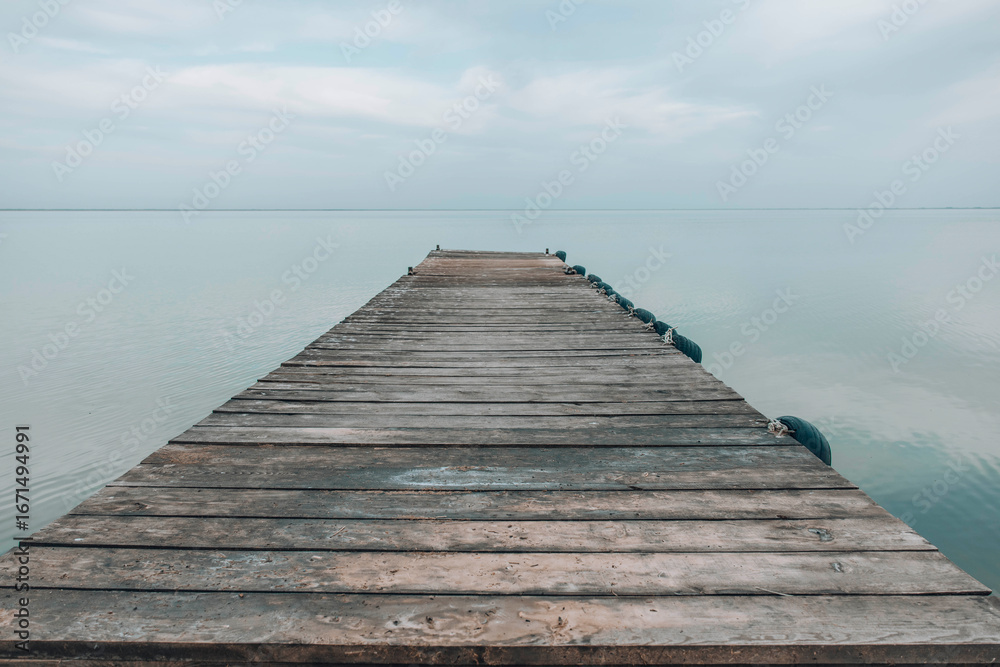 Fototapeta premium wooden pier stretching into calm still lake under soft cloudy sky, peaceful waterscape scene with muted colors and quiet atmosphere, travel inspiration, nature therapy, wellness retreats