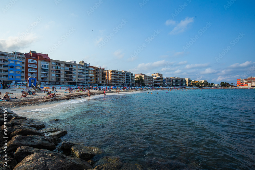 Fototapeta premium A panoramic view of Torrevieja’s coastline, showing a curved sandy beach lined with apartment buildings and the calm Mediterranean Sea. The scene captures a lively seaside atmosphere with people relax