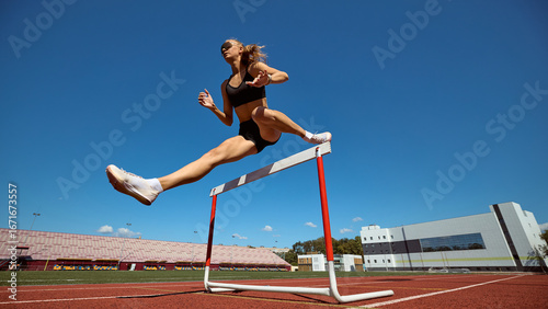 Bild auf Leinwand Female hurdler mid-air over hurdle with dynamic stride on stadium track