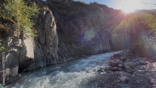 Beautiful landscape with a mountain river flowing among rocky cliffs. Sunlight shines in the background, creating a scenic wilderness view with natural water stream, rugged terrain, and peaceful outdo