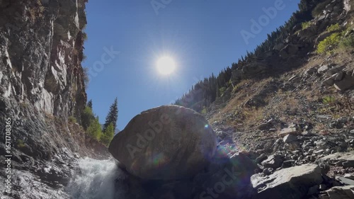 Beautiful mountain river flows from a huge stone surrounded by rocky cliffs and coniferous forest. Scenic wilderness view with natural water stream, mountain landscape, and peaceful outdoor atmosphere