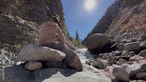 Man sits on a huge stone against the backdrop of a beautiful mountain landscape. Scenic view with mountain river, rocky terrain, and wilderness nature creating peaceful outdoor atmosphere of travel, e
