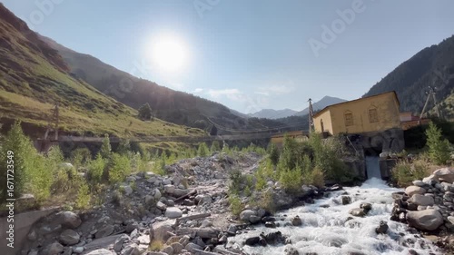 Beautiful mountain landscape with a hydroelectric power station building standing on a mountain river. Sunlight shines over the peaks in the background, combining industrial architecture with natural 