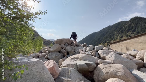 Man in black clothes with backpack climbs over a huge pile of rocks in the mountains. He stops to admire scenic natural beauty with rocky slopes, cloudy sky, and wilderness landscape in highland terra