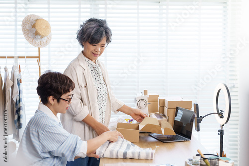 Senior asian mother with short gray hair and adult asian daughter working together in fashion atelier small business packing clothing into cardboard box for online shop shipping, business teamwork