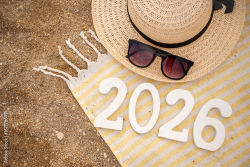 Stylish sunglasses and a fashionable straw hat resting on a striped beach towel with wooden numbers 2026, set against golden sand, evoke anticipation for a new year's beach vacation