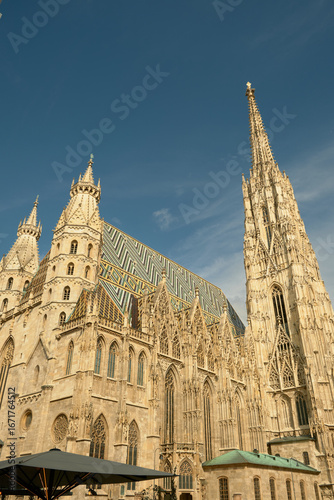 Wide view of St. Stephen’s Cathedral with its Gothic spires and mosaic roof, framed by a café umbrella in the foreground, under a bright sunny sky.