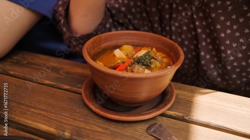 A person savors a bowl of lagman with beef featuring colorful vegetables while seated at a rustic wooden table in a charming restaurant during midday