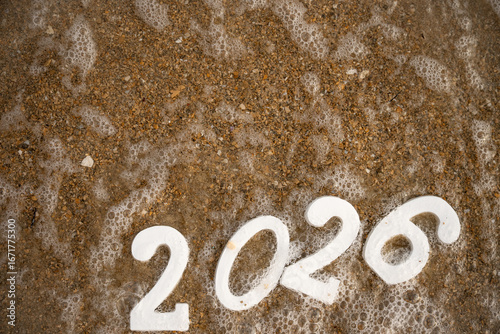 White numbers 2026, partially covered by sea foam, rest on a sandy beach, embodying the spirit of celebration for the upcoming new year while capturing a serene coastal scene