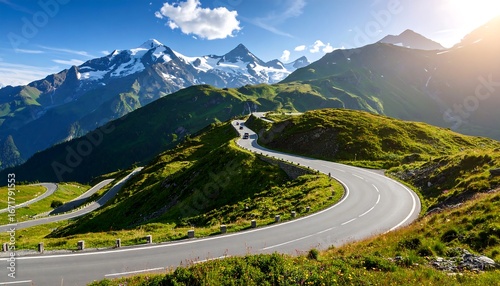 A winding mountain road ascends through lush green meadows, framed by majestic snow-capped peaks under a brilliant blue sky.