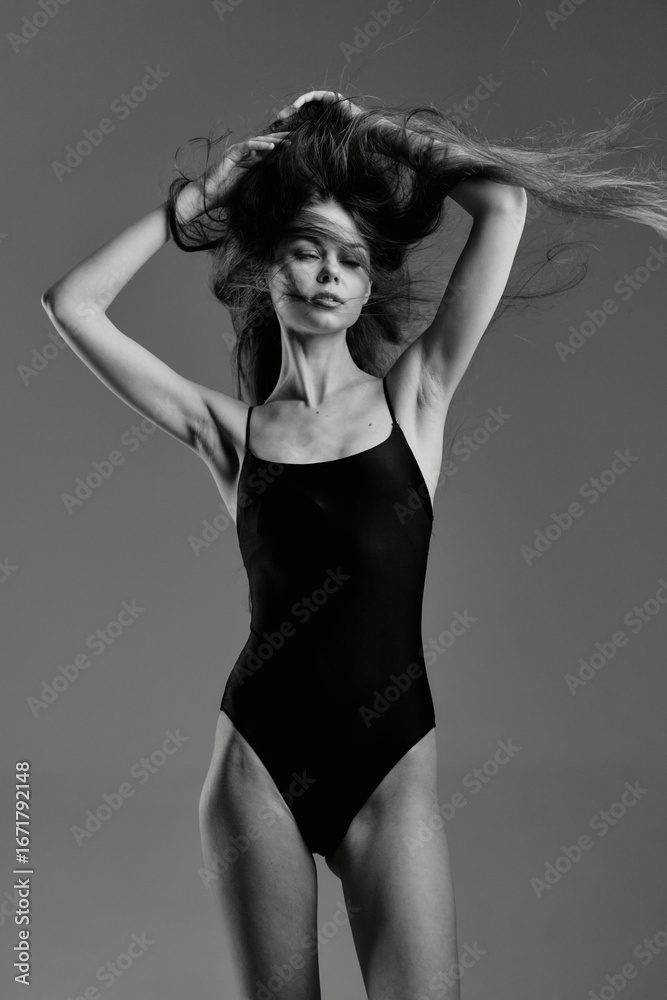 Fototapeta premium Black and white portrait of a slender woman in a black swimsuit with flowing hair on a gray background.