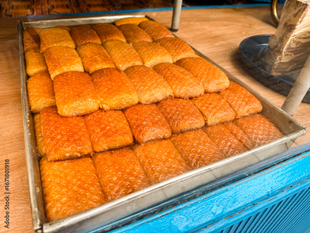 Fototapeta premium Traditional sweet pastries displayed on metal tray with golden syrup surface in bakery shop. Food culture, dessert preparation, confectionery tradition and culinary presentation.