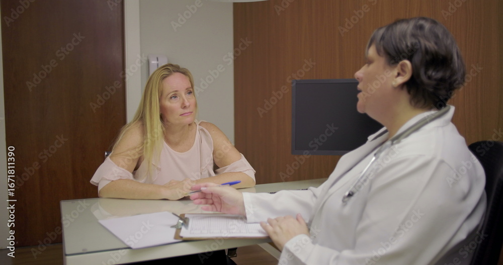 Obraz premium Middle-aged woman patient listening to doctor during medical consultation at desk in clinical office setting with clipboard and documents