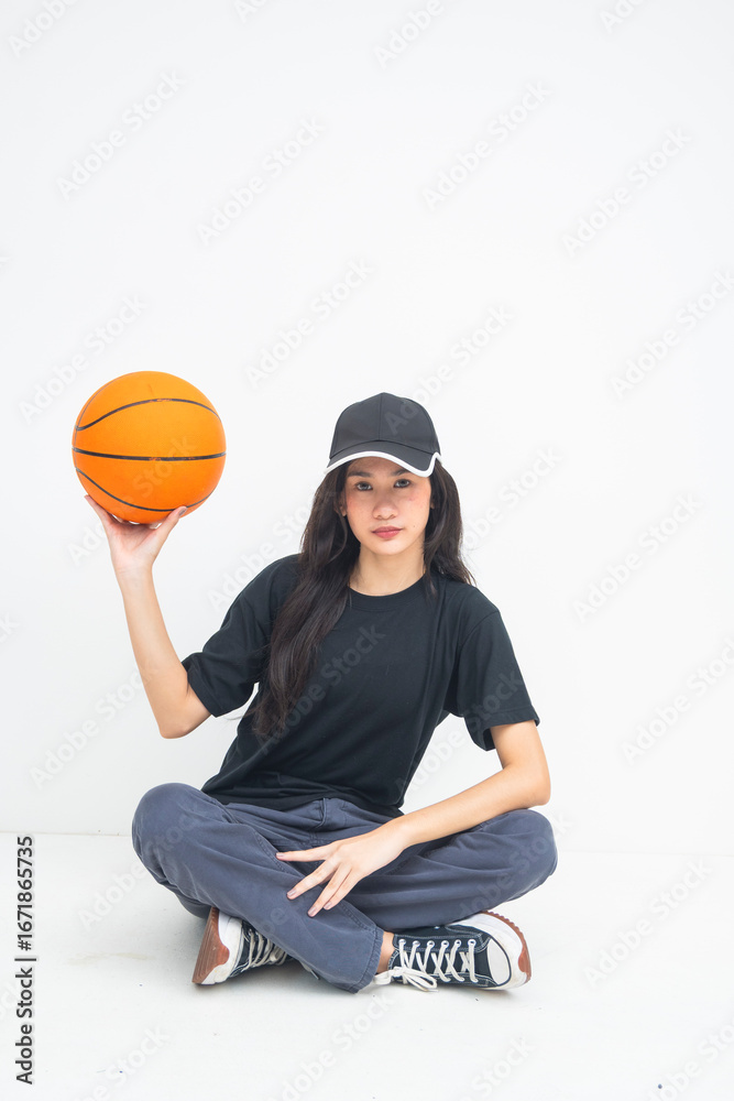 Fototapeta premium A confident young Asian woman captured sitting cross-legged with a basketball in her hands, showing strength and a fierce, empowered demeanor. Isolated on a white background.