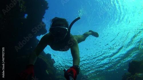 selfie of a female free diver wearing bikini snorkel and mask but with no fin just bare foot diving swimming down into ocean underwater surrounds by coral rock reef with other people on top of water