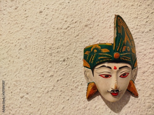 A traditional Indonesian mask with a striking expression hangs on a textured white wall.