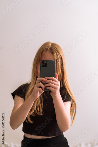 Young girl taking a selfie with a smartphone, standing in front of a light wall. Minimalist and candid lifestyle moment with soft natural lighting.
