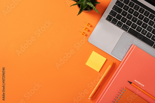 Laptop keyboard with orange stationery, notebook, pen, and sticky note on desk. Workplace with copyspace.