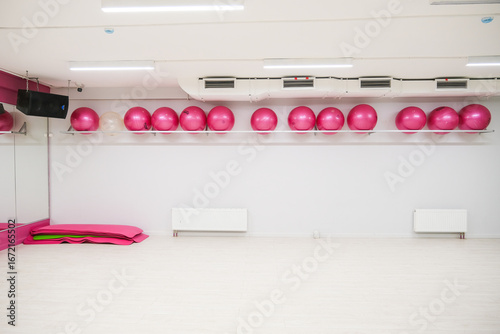 Many pink exercise balls and yoga mats stored in an empty gym. Fitness studio interior with clean white walls and light wood floor for sports activity.