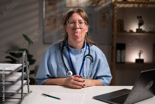 фотографія nurse working overtime on night shift showing tongue and looking at the camera in hospital office