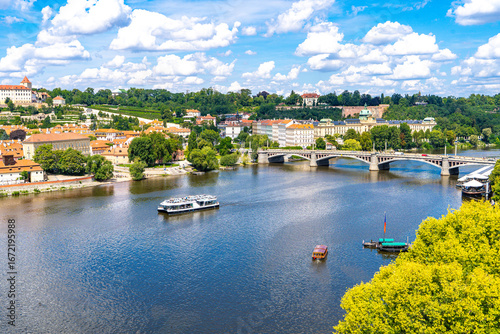 Photography he Vltava River, surrounded by lush summer greenery, with a bridge connecting the historic districts of the Old Town under a blue sky with clouds