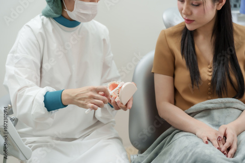 Asian female dentist using toothbrush on dental model to show correct brushing technique to adult asian female patient during oral health consultation in dental clinic after orthodontic procedure
