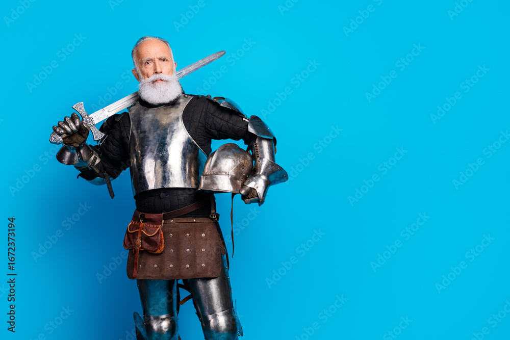 Obraz premium Portrait of a senior man in full medieval knight armor holding a sword and helmet with bright blue background