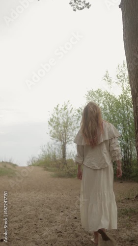 A girl walks along a sandy path in a beautiful white dress, surrounded by serene, breathtaking nature