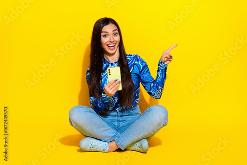Attractive woman sitting cross-legged on yellow background holding smartphone in hand and pointing energetically to side.