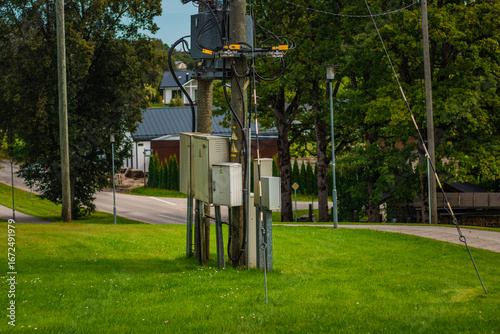 A utility pole equipped with electrical boxes and cables stands in a grassy suburban area near a quiet street and houses.