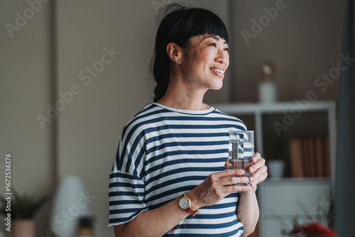 Smiling japanese woman drinking water and looking out the window at home