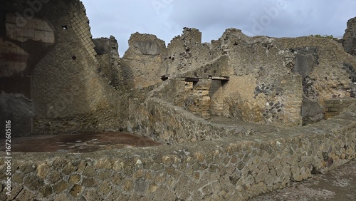 Herculaneum, Italy - 9 January 2025. Stone ruins of Roman-era buildings with mosaic-patterned walls and remains of entrances and passageways under cloudy skies.