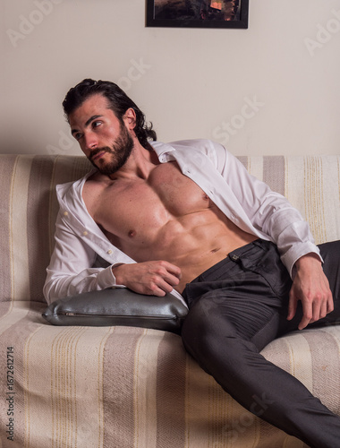 A man reclines casually on a striped sofa, wearing an unbuttoned white shirt and black trousers. He looks pensive, with a relaxed yet stylish demeanor, set in a cozy room