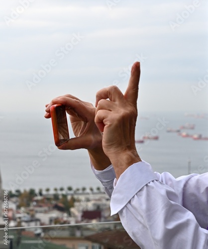 Person holding smartphone, capturing scenic view of ocean and ships, with fingers forming a frame, showcasing photography and creativity in urban environment