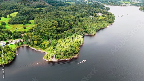 aerial high angle drone shot of Ullswater lake with forrest trees on lakeside panning right over nature in summer with sun peeking through clouded skies