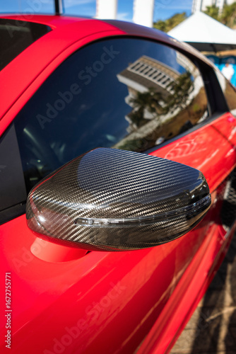 Car side mirror with a carbon fiber cover and integrated turn signal repeater, on a vehicle with red paint.