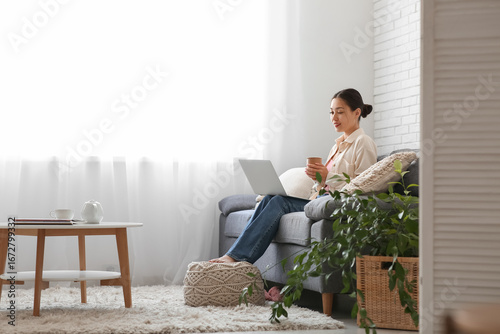Tableau sur toile Young Asian woman using laptop on sofa near light curtains at home
