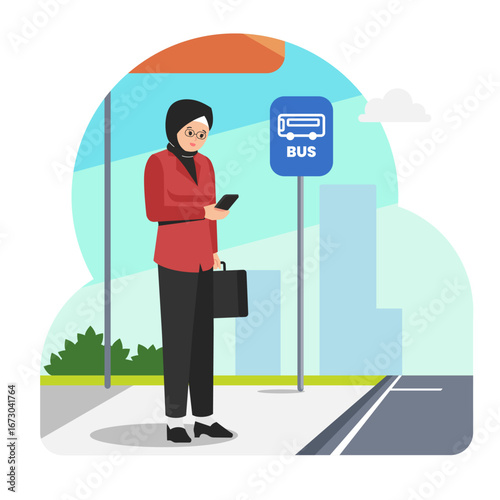 female office worker wearing a hijab waits for a bus at a public bus station while using her smartphone.