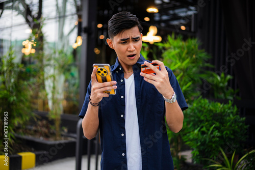 Surprised young Asian man in navy shirt and white t-shirt looks at his declined credit card and smartphone in disbelief. Online payment refused or blocked card.