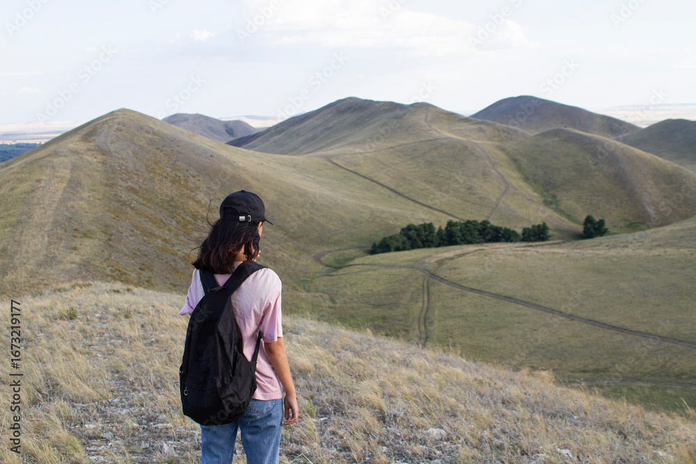 Naklejka premium a teenage girl is standing with her back to the camera, holding a black backpack and looking out at the Karamuruntau mountain range