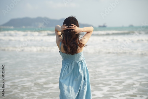 A woman in a light blue dress stands on the beach, facing the sea, with her hands on her head, enjoying the seaside view.