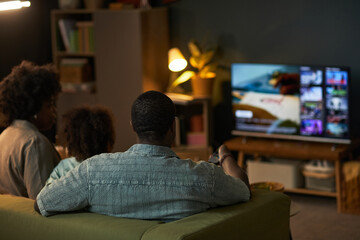 Black man, Black woman, and child sitting on sofa watching television together in living room, remote control in man's hand, family spending leisure time