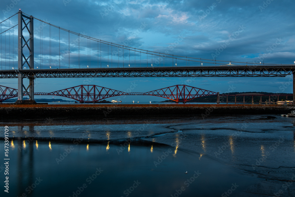 Naklejka premium Taken at dusk, a view down the Firth of Forth from Queensferry, with dark blue sky and reflections in the water.