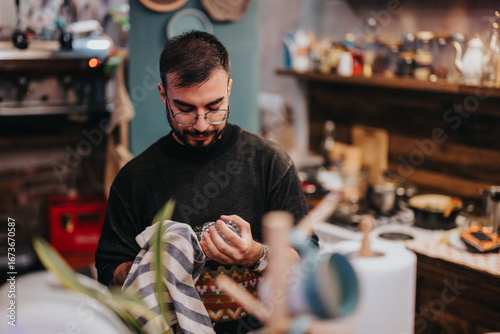 Φωτογραφία A young man wearing glasses and cleaning a glass with a striped cloth in a cozy, rustic cafe-like environment, showcasing warmth and attention to detail