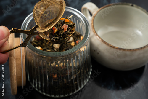 Fotografie A hand holds a metal infuser above a glass jar filled with loose herbal tea leaves, while a simple teapot sits nearby