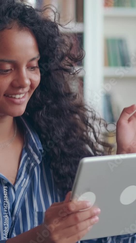 Young Indian woman with curly hair is engaged in cheerful online conversation, smiling and waving at camera. She sits in cozy indoor space filled with books and plants, exuding warmth and positivity