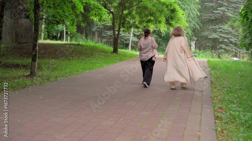Two friends enjoy a walk through a lush green park on a pleasant day while wearing casual outfits and a light cape