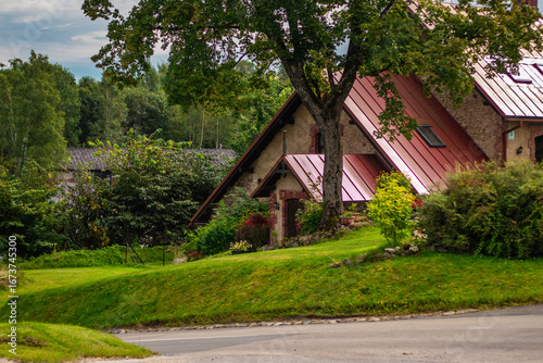 A cozy countryside home with steep red metal roofs sits among lush green trees and garden plants beside a quiet rural road.