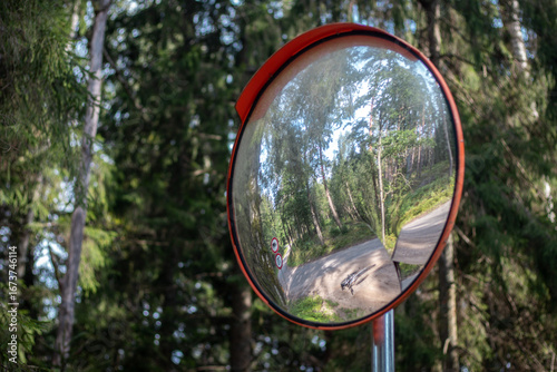 Fototapeta A roadside convex mirror reflects a curving forest road with trees, a bicycle, and multiple traffic signs in natural daylight